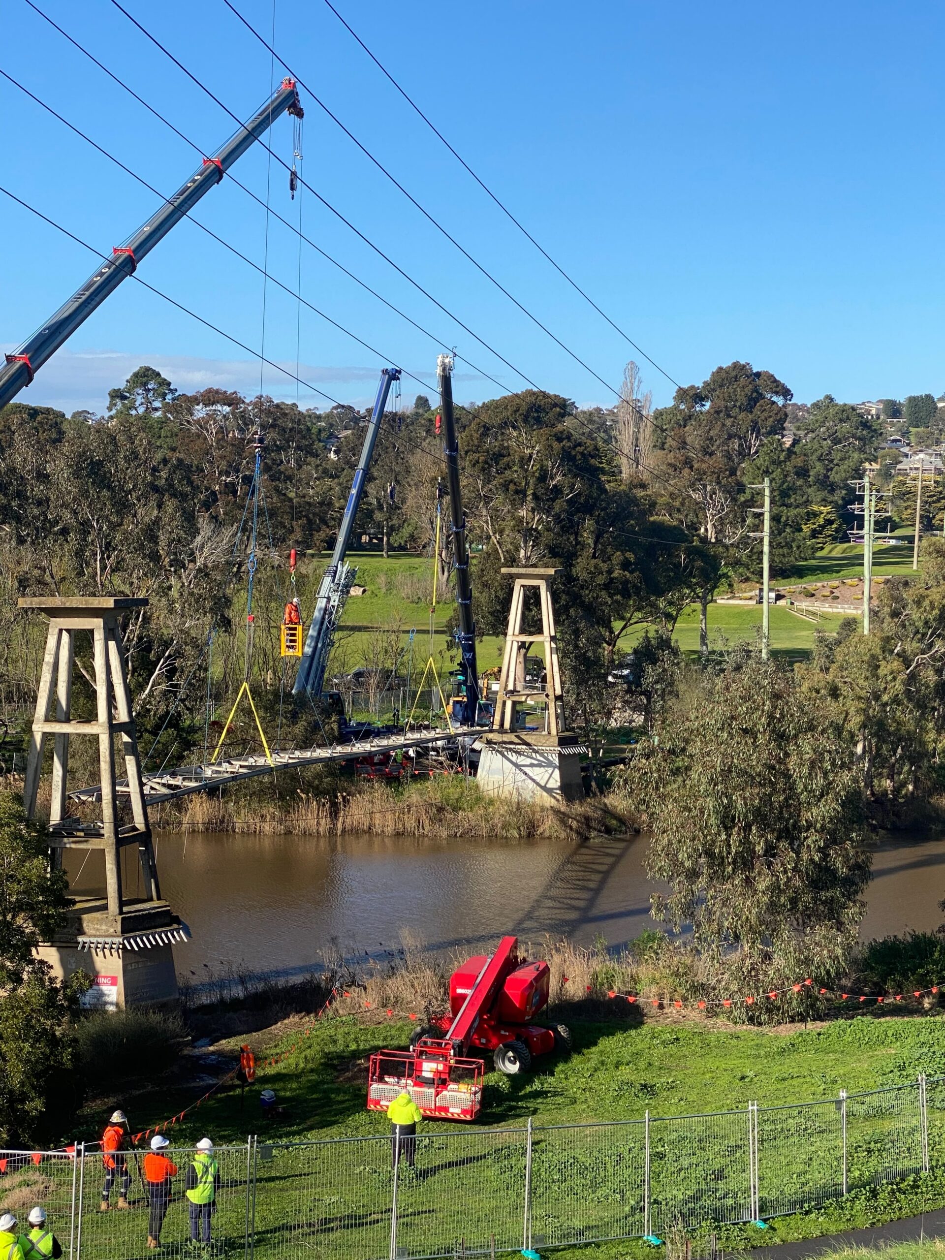 Barwon River suspension bridge deck rigged to cranes on both banks between the concrete pylons, with EWP and Digga crew on the north bank, Fyansford